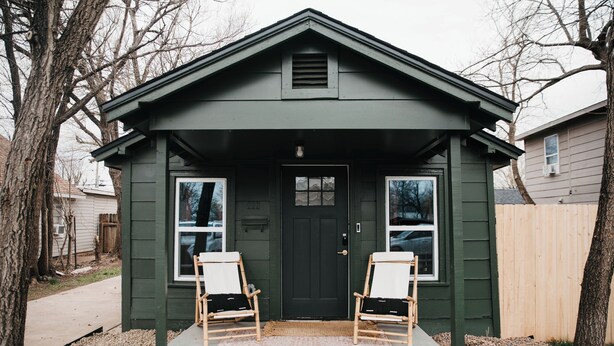 A small, green wooden building with a dark door, two windows, and two wooden chairs in front.