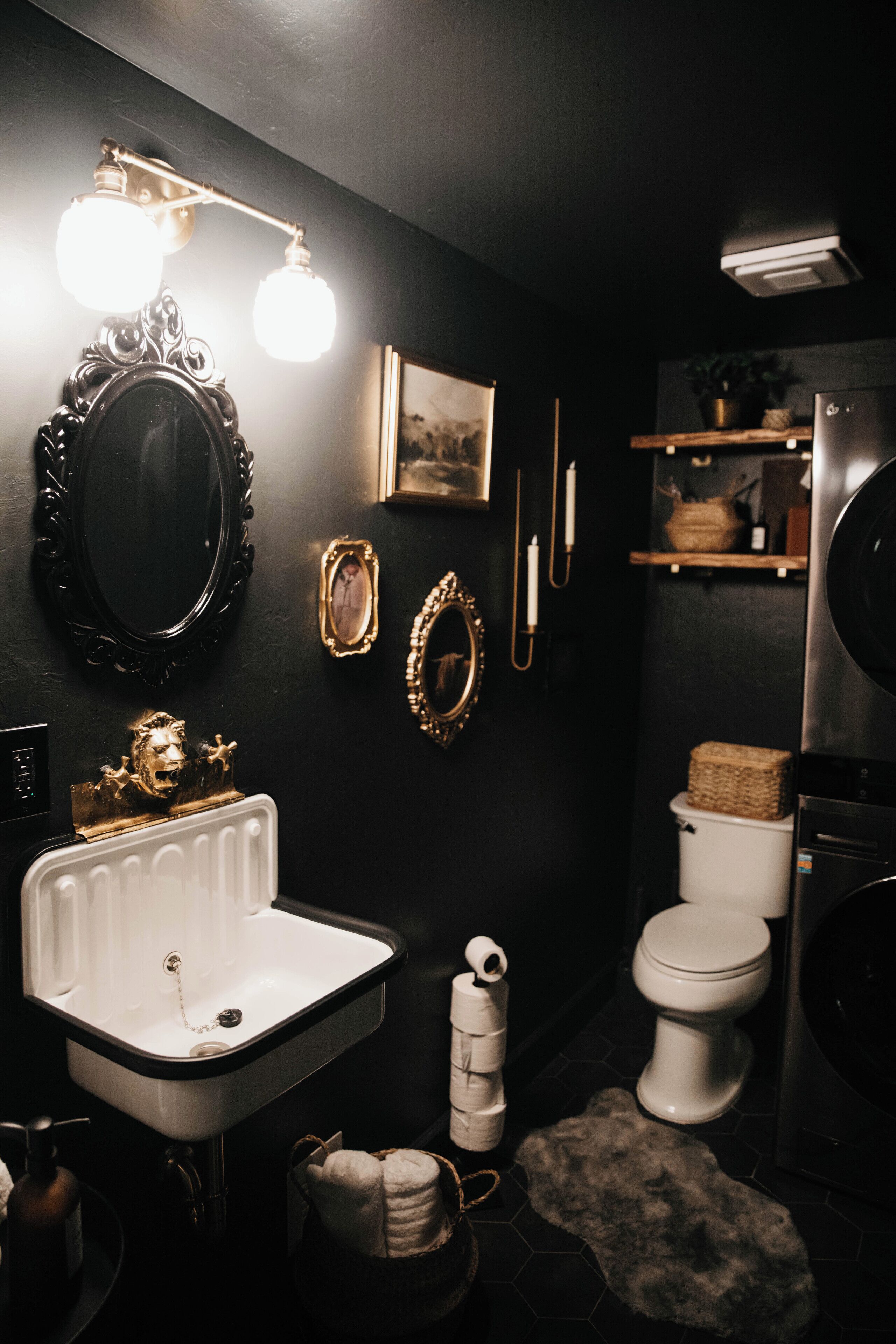 Un baño con lavamanos blanco, paredes negras y un inodoro blanco. Hay espejos decorativos y una lámpara sobre el lavamanos.