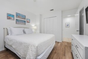 A bedroom with a large bed, white walls, and wooden flooring. There are framed pictures on the wall and a white dresser with drawers. - Perfect Getaway – Beach Access, Pool & Hot Tub (Destin)