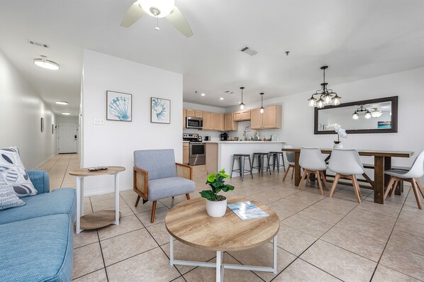 A modern living room with a blue sofa, a round coffee table, a small side table, a grey armchair, and a dining area with a wooden table and chairs. - Perfect Getaway – Beach Access, Pool & Hot Tub (Destin)
