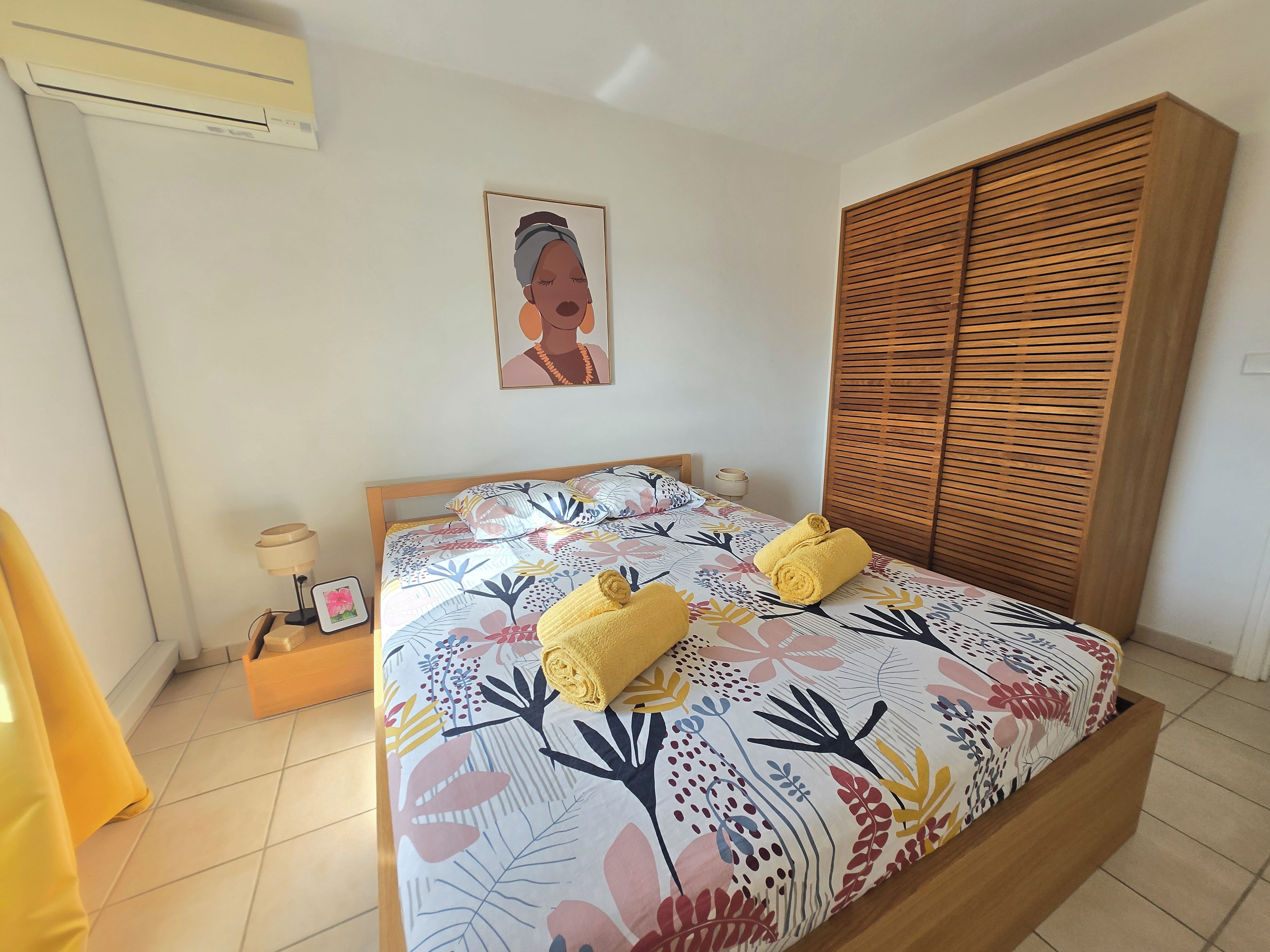 A bedroom with a bed, a wooden wardrobe, a bedside table with a lamp, and a framed picture on the wall.