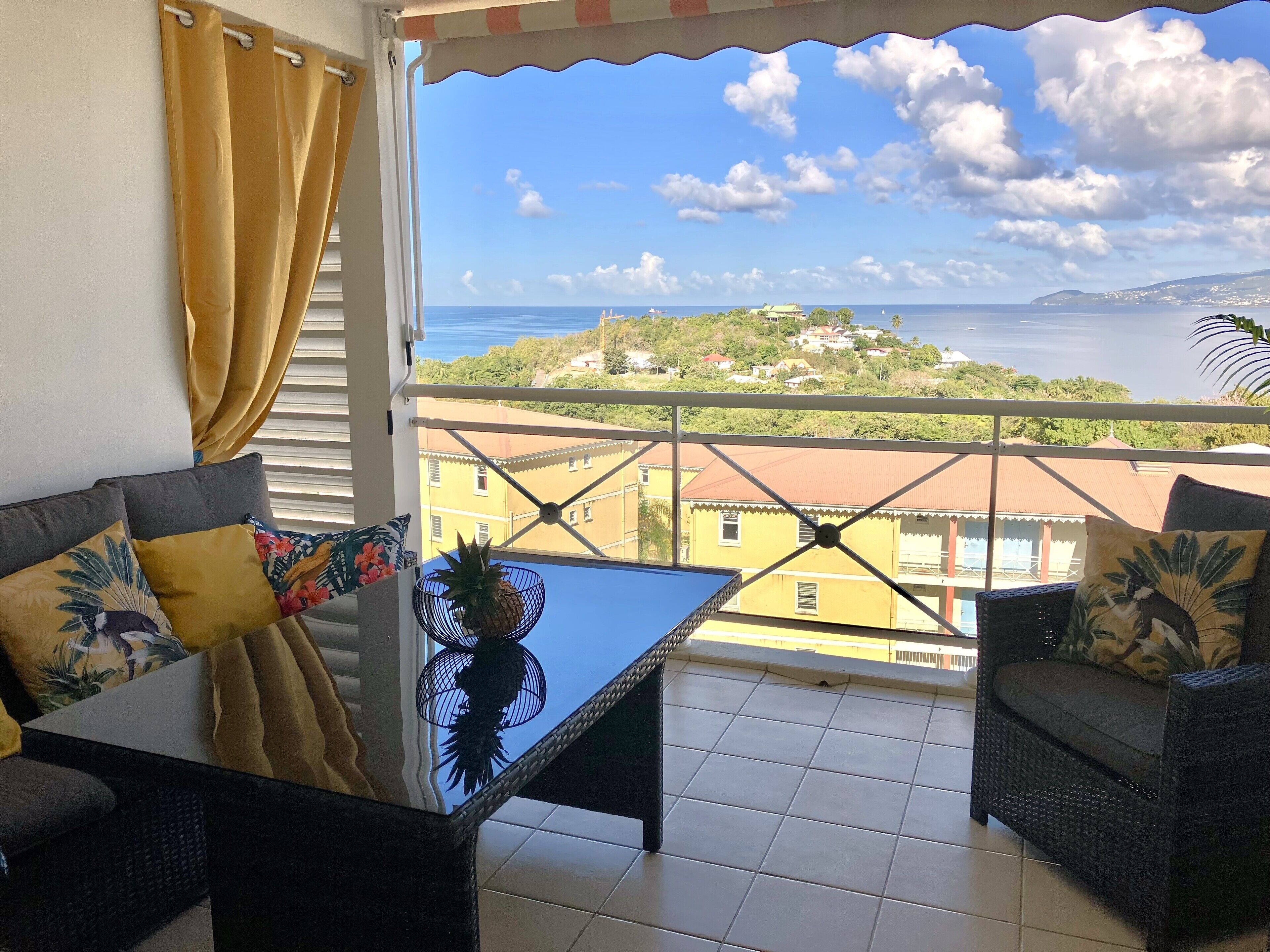 Un balcón con vista al mar y edificios. Hay una mesa con una piña y un cuenco decorativo. Dos sillas de mimbre con cojines están en el balcón.