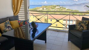 Uma varanda com vista para o mar e edifícios. Há uma mesa com uma abacaxi e uma tigela decorativa. Duas cadeiras de vime com almofadas estão na varanda.