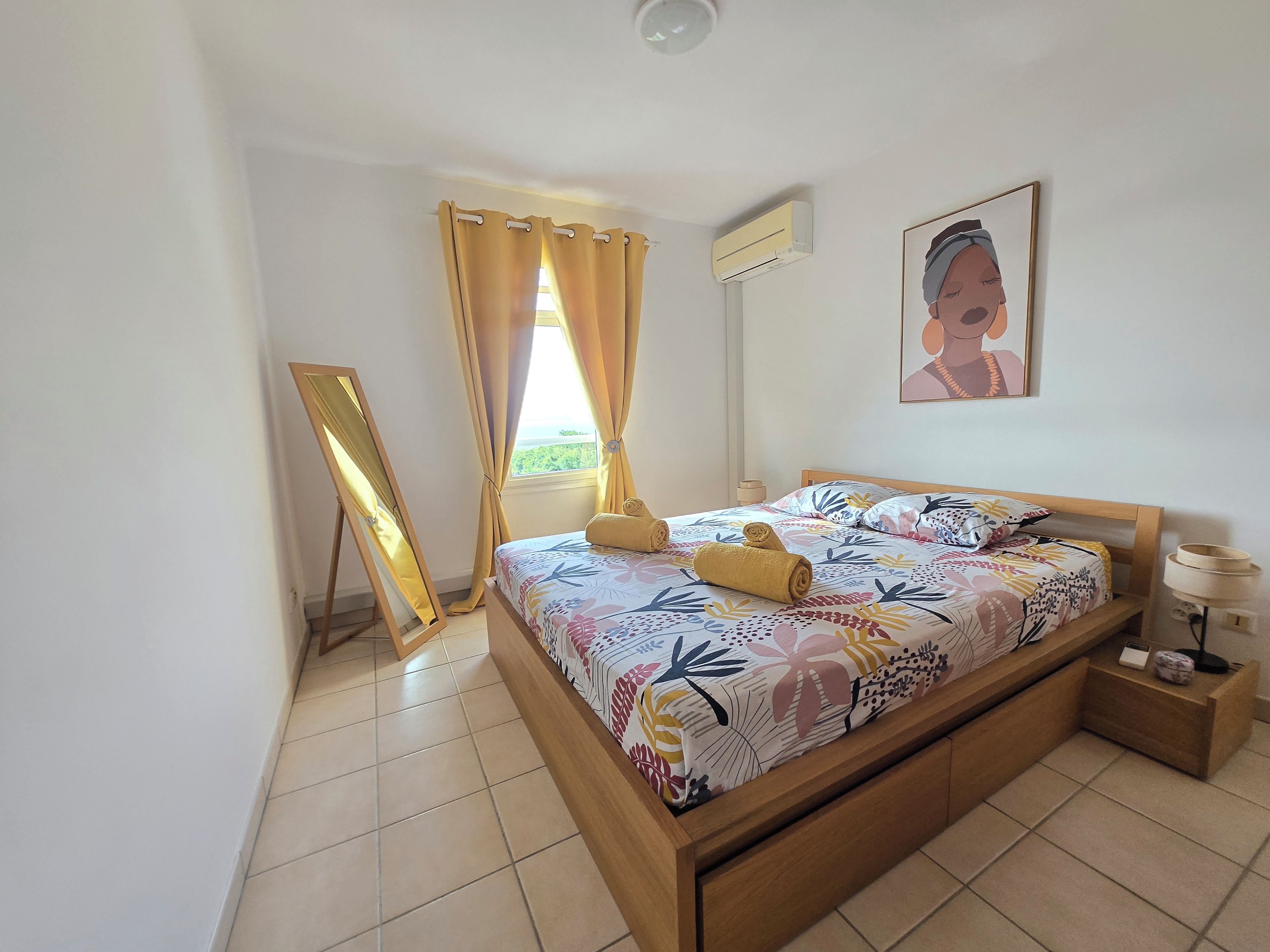 Une chambre à coucher comprenant un lit, une table de chevet, un miroir, une fenêtre avec des rideaux et un appareil de climatisation.