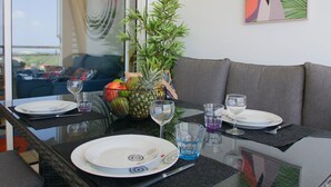 Uma área de refeições com uma mesa de vidro posta para dois, com pratos, talheres e copos. Há uma fruteira com abacaxi e manga sobre a mesa, e uma planta em vaso ao fundo.