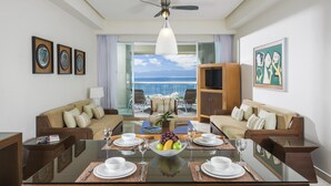 Un coin repas avec une table en verre, des chaises et une vue sur l’océan. On y trouve également des canapés et un téléviseur fixé au mur.
