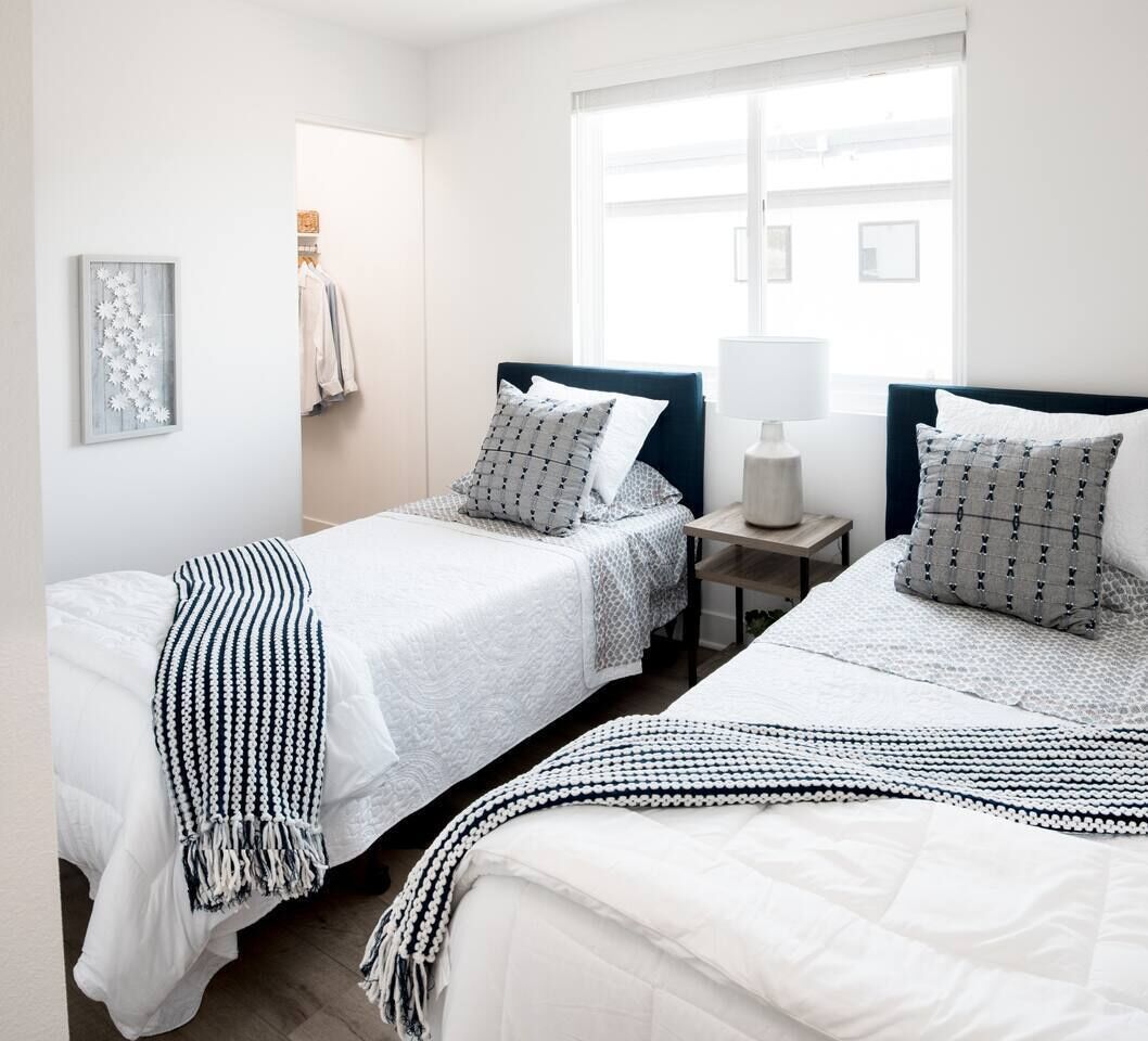 A hotel room with two beds, a bedside table with a lamp, a window with a view, and a framed picture on the wall.