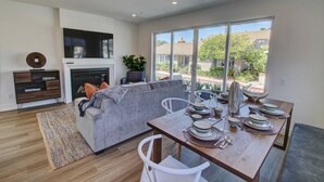 Una sala de estar moderna con un sofá gris, una mesa de comedor con platos y vasos, una chimenea y ventanas amplias que ofrecen vistas del vecindario.