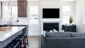 Eine moderne Küche mit einer zentralen Kochinsel, Barhocker, einem Kamin und einem großen, grauen Sofa im Wohnbereich.
