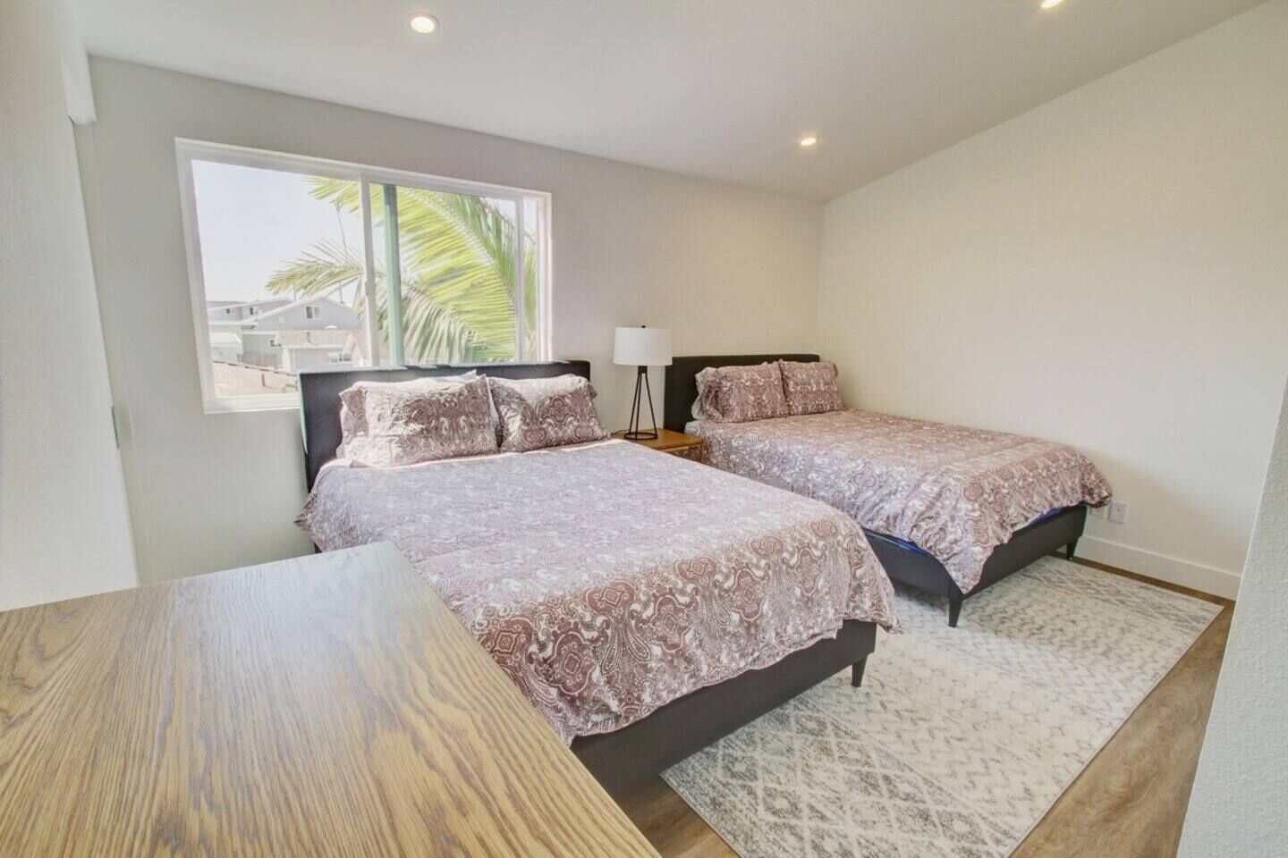 A bedroom with two beds, a wooden table, and a window with a view of palm trees.