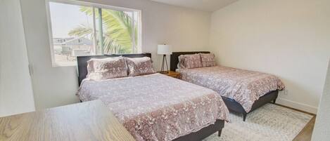 A bedroom with two beds, a wooden table, and a window with a view of palm trees.