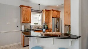 A kitchen with wooden cabinets, a stainless steel refrigerator, and a black countertop island with stools. - 8min Texas A&M, 1 King Beds, firepit, GriddleGrill (College Station)