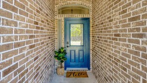 A narrow brick corridor leading to a blue door with a small potted plant beside it and a 'Hello' mat at the entrance. - 8min Texas A&M, 1 King Beds, firepit, GriddleGrill (College Station)