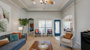 A spacious living room with a ceiling fan, a blue sofa, a coffee table, and a door leading to another room. - 8min Texas A&M, 1 King Beds, firepit, GriddleGrill (College Station)