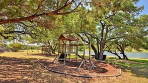 Une aire de jeux avec des balançoires et un petit abri, entourée d’arbres et bordée d’un lac.
