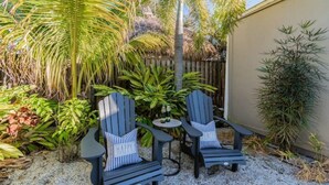 Twee Adirondack stoelen met kussens, een klein rond tafeltje met flessen en diverse tropische planten in een tuin.