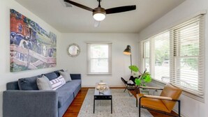 A living room with a gray sofa, a coffee table, a ceiling fan, a window with blinds, and a painting on the wall. - River, Wine, Hot Tub, 2 King Beds, Large backyard (Blanco)