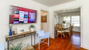 A modern living room with a flat-screen TV displaying various streaming services, a dining area with wooden furniture, and a hallway leading to another room. - River, Wine, Hot Tub, 2 King Beds, Large backyard (Blanco)