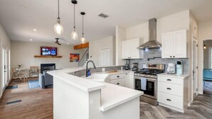A modern kitchen with white cabinets, a central island, stainless steel appliances, pendant lighting, and a visible TV in the background. - 5 kings, 3 acres, lake view, cabana, firepit/stars (Buchanan Dam)