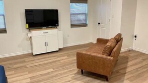 Una sala de estar con una televisión de pantalla plana sobre un mueble blanco, un sofá marrón y piso de madera.
