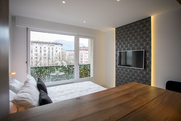 Un dormitorio moderno con un ventanal grande, una cama, una mesa de madera y un televisor montado en la pared.