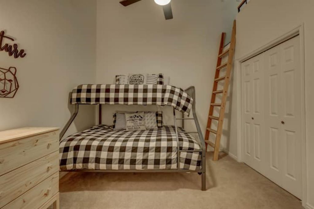 A bunk bed with checkered bedding, a wooden ladder, a dresser, and a closet with white doors.