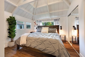 A bedroom with a large bed, a potted plant, a wooden bench, and a view of greenery through the window.