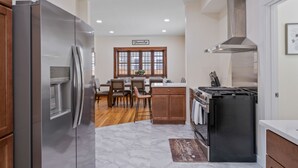 A modern kitchen with stainless steel appliances, wooden cabinets, a dining area with chairs, and a large window. - Eastown Luxe (Grand Rapids)