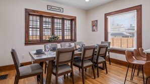A dining area with a wooden table, grey upholstered chairs, and a window with blinds. - Eastown Luxe (Grand Rapids)
