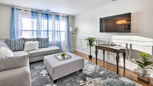 A living room with a grey sofa, patterned cushions, a coffee table with a tray, a flat-screen TV mounted on the wall, a wooden side table, and a potted plant. - Bay Shore Vacation Rental w/ Shared Yard! (Bay Shore)