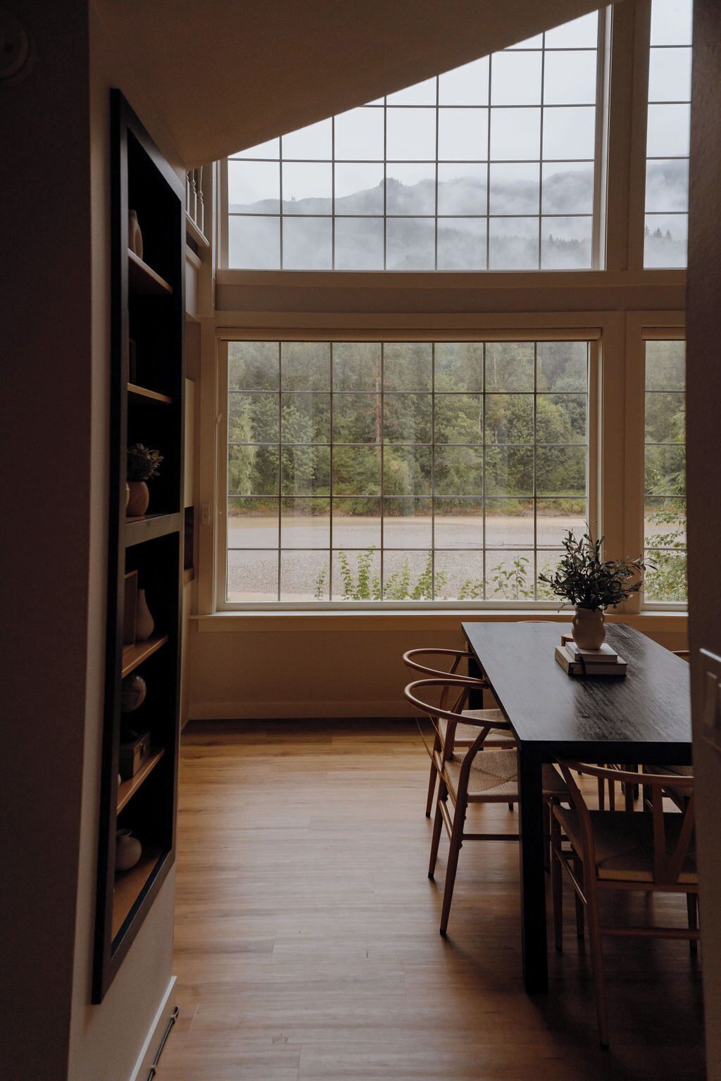 A dining area with a table and chairs, a bookshelf, and a large window with a view of a forest and a road.