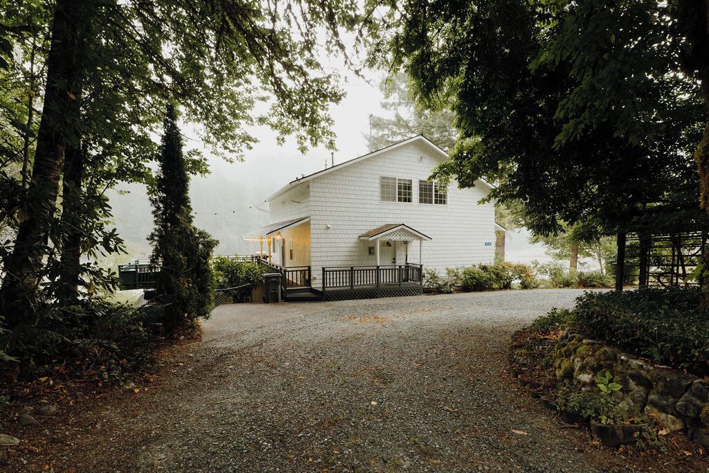 A white two-story house with a porch, surrounded by trees and a gravel driveway.