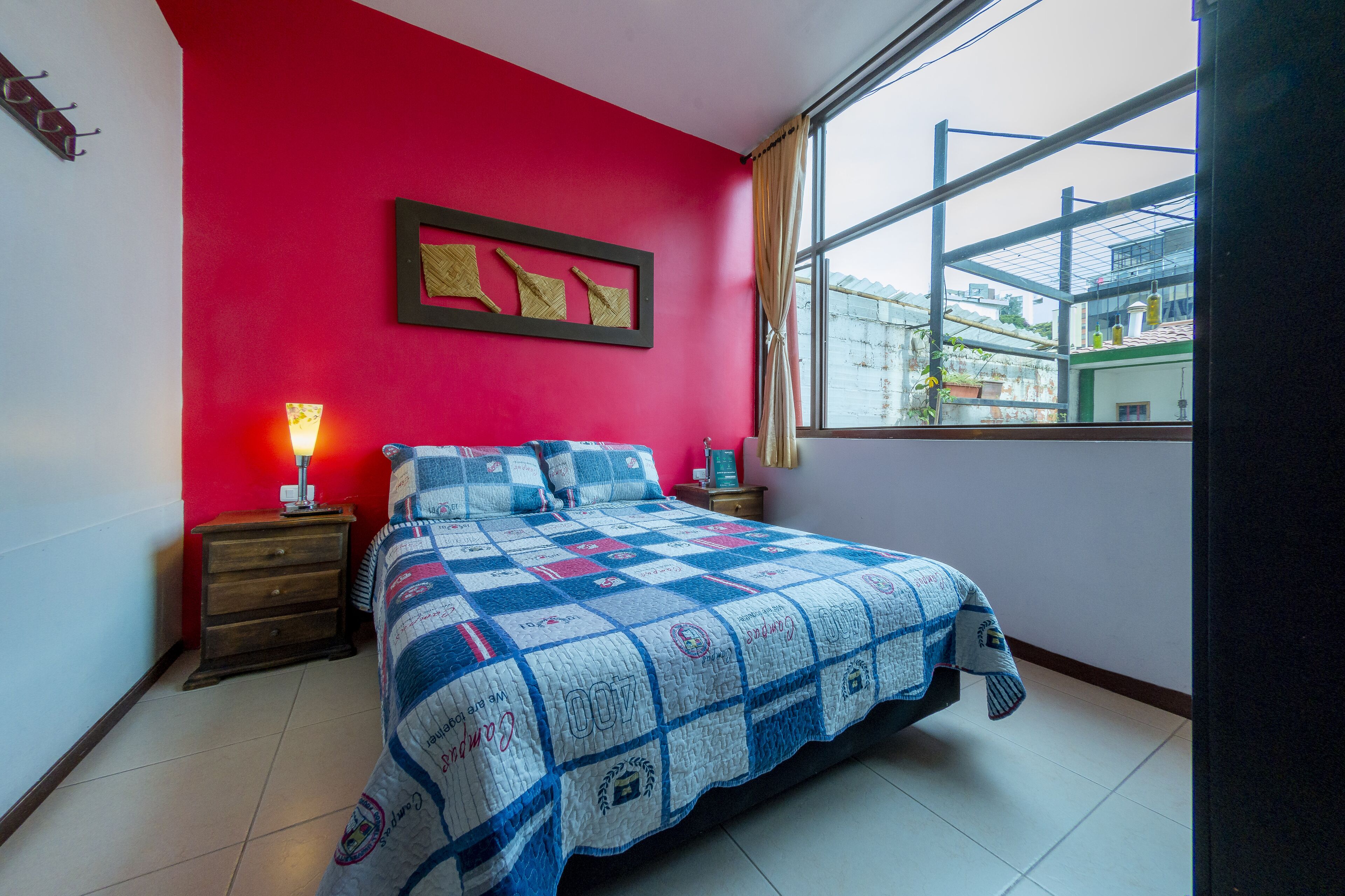 A bedroom with a red wall, a large bed with a quilt, a nightstand with a lamp, and a window with a view of buildings.