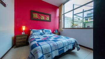A bedroom with a red wall, a large bed with a quilt, a nightstand with a lamp, and a window with a view of buildings.