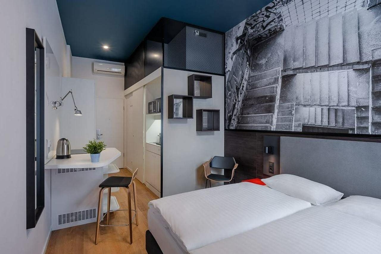 Quarto de hotel moderno com cama, escrivaninha e cadeira. Há um ar condicionado fixado na parede e uma pequena planta na escrivaninha.