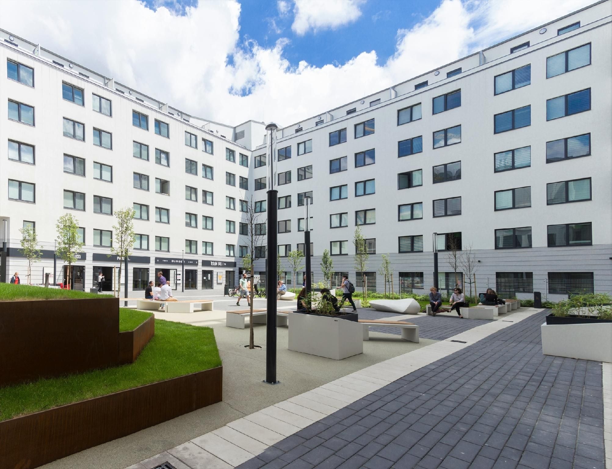 Un bâtiment moderne à plusieurs étages, doté de grandes fenêtres et d’une cour intérieure aménagée avec des espaces de repos et de la verdure.
