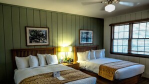 Une chambre d’hôtel avec deux lits, des têtes de lit en bois, un mur vert, des tableaux encadrés, une fenêtre avec des stores et un ventilateur de plafond.