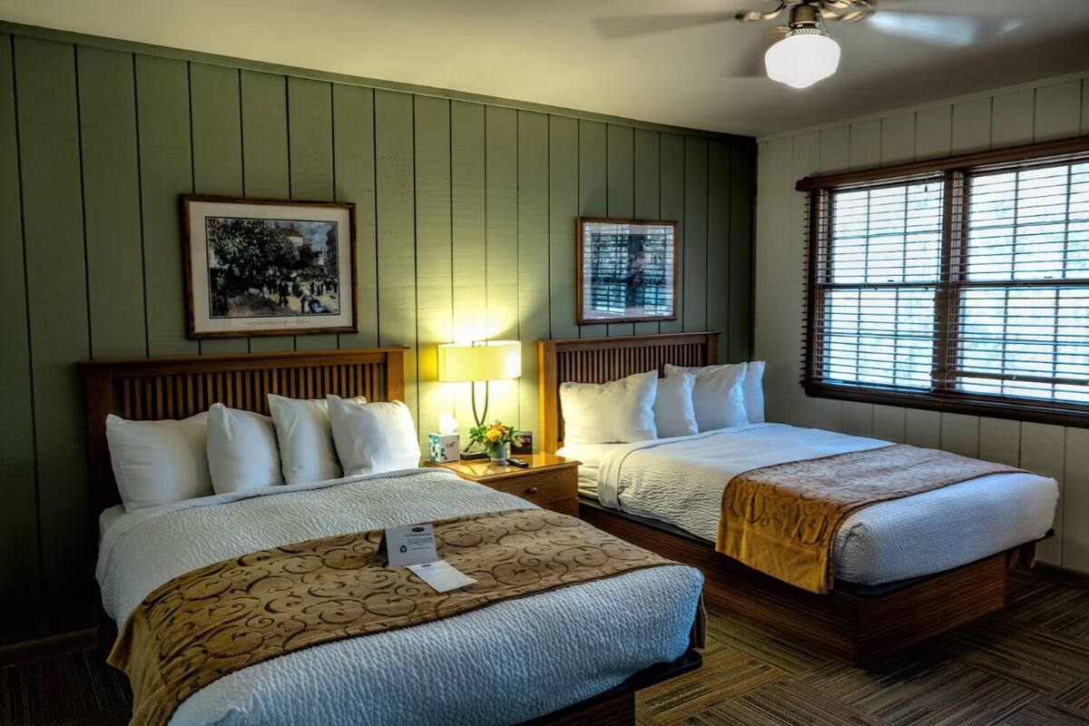 A hotel room with two beds, wooden headboards, a green wall, framed pictures, a window with blinds, and a ceiling fan.