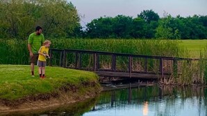 Un plan d’eau paisible, surmonté d’un pont en bois, entouré de verdure et sous un ciel dégagé.