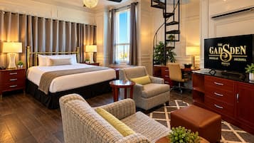A hotel room with a king-sized bed, two bedside tables, a desk, a chair, a sofa, a coffee table, a TV, a ceiling fan, a window with curtains, and a plant.