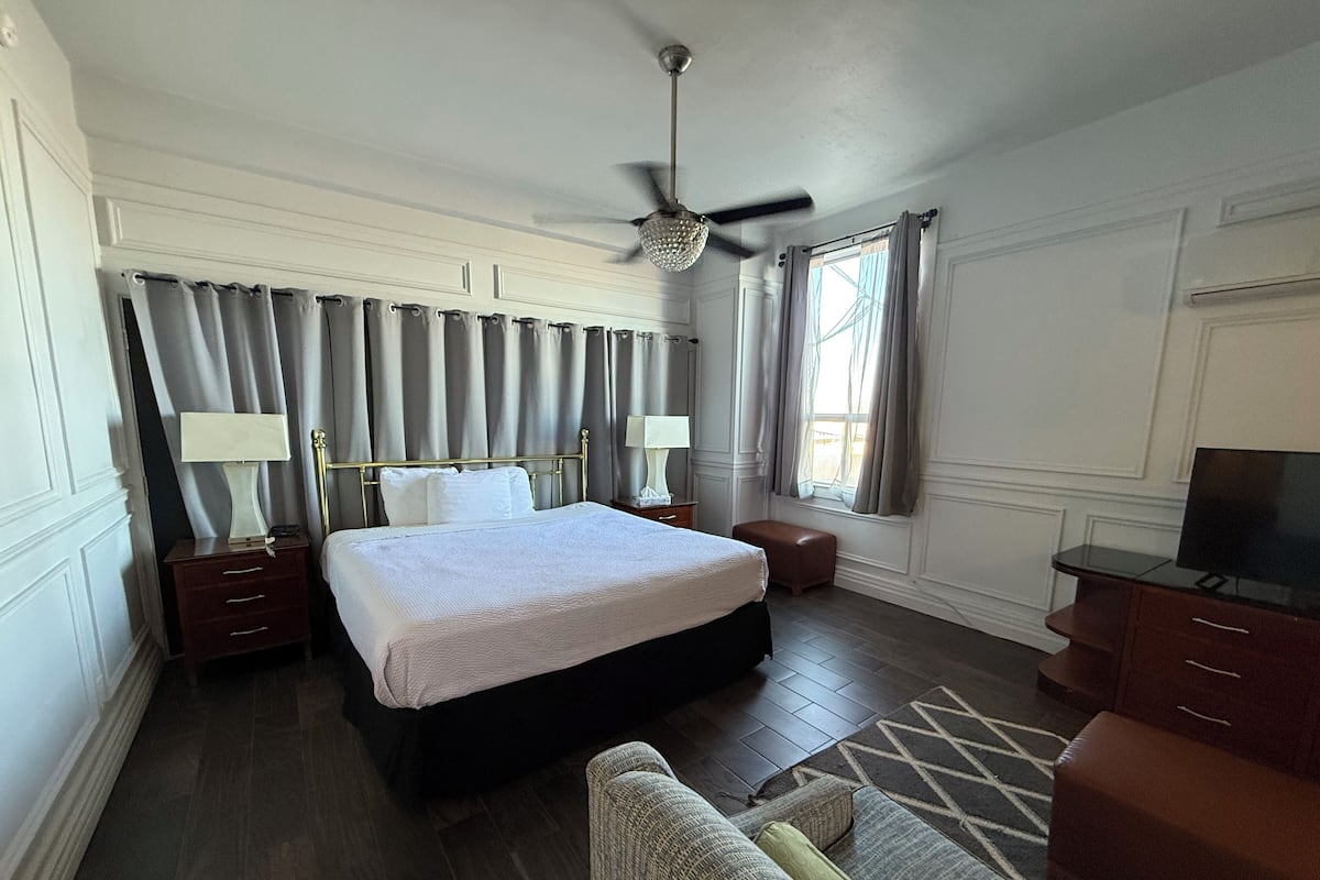 Een hotelkamer met een groot bed, een plafondventilator, een televisie, een bureau en een raam met gordijnen.