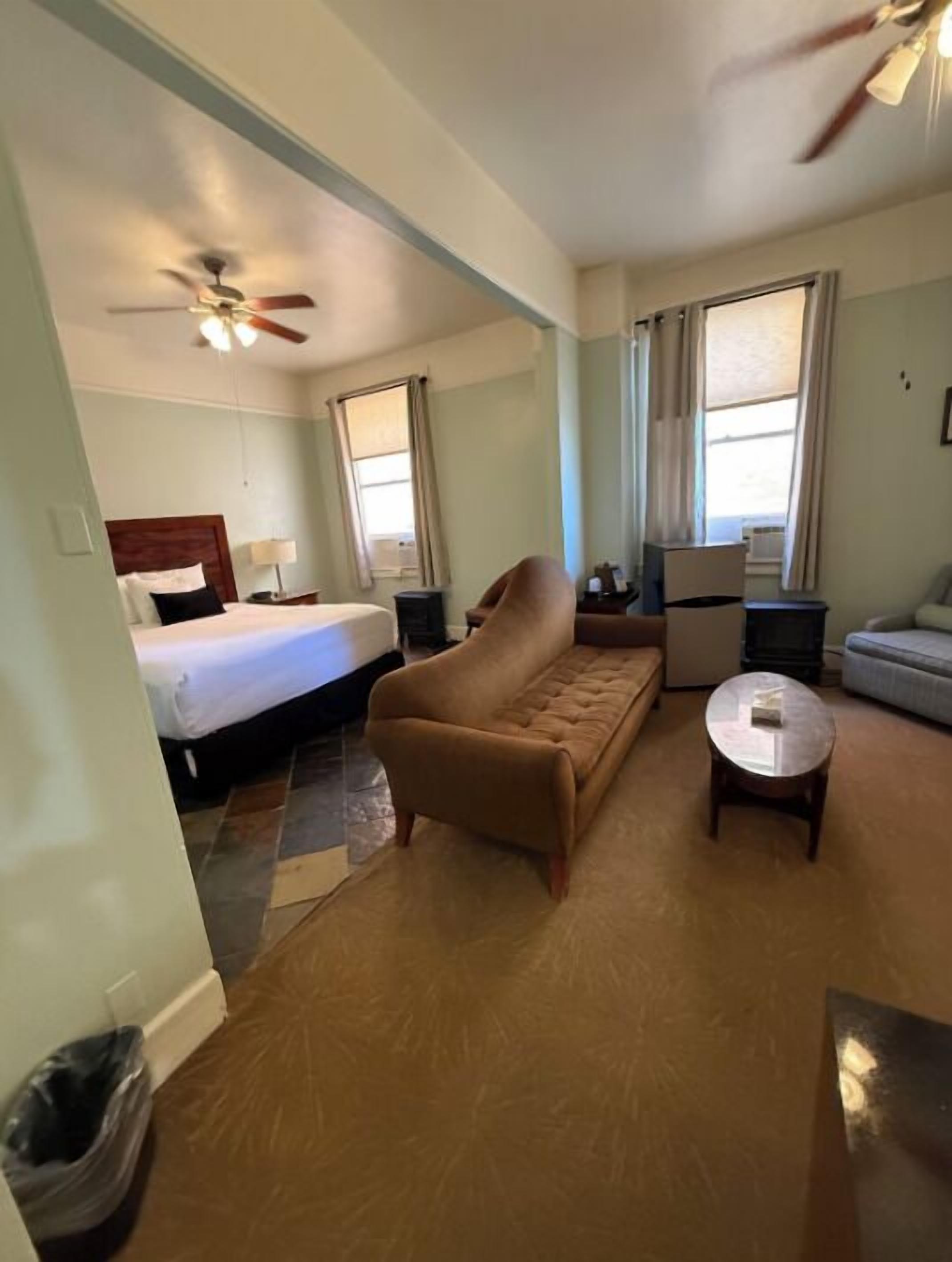 A hotel room with a bed, a sofa, a small table, a ceiling fan, and windows with curtains.
