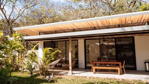 Een modern huis met een overdekte patio, grote ramen en een tuin met planten en een bankje.