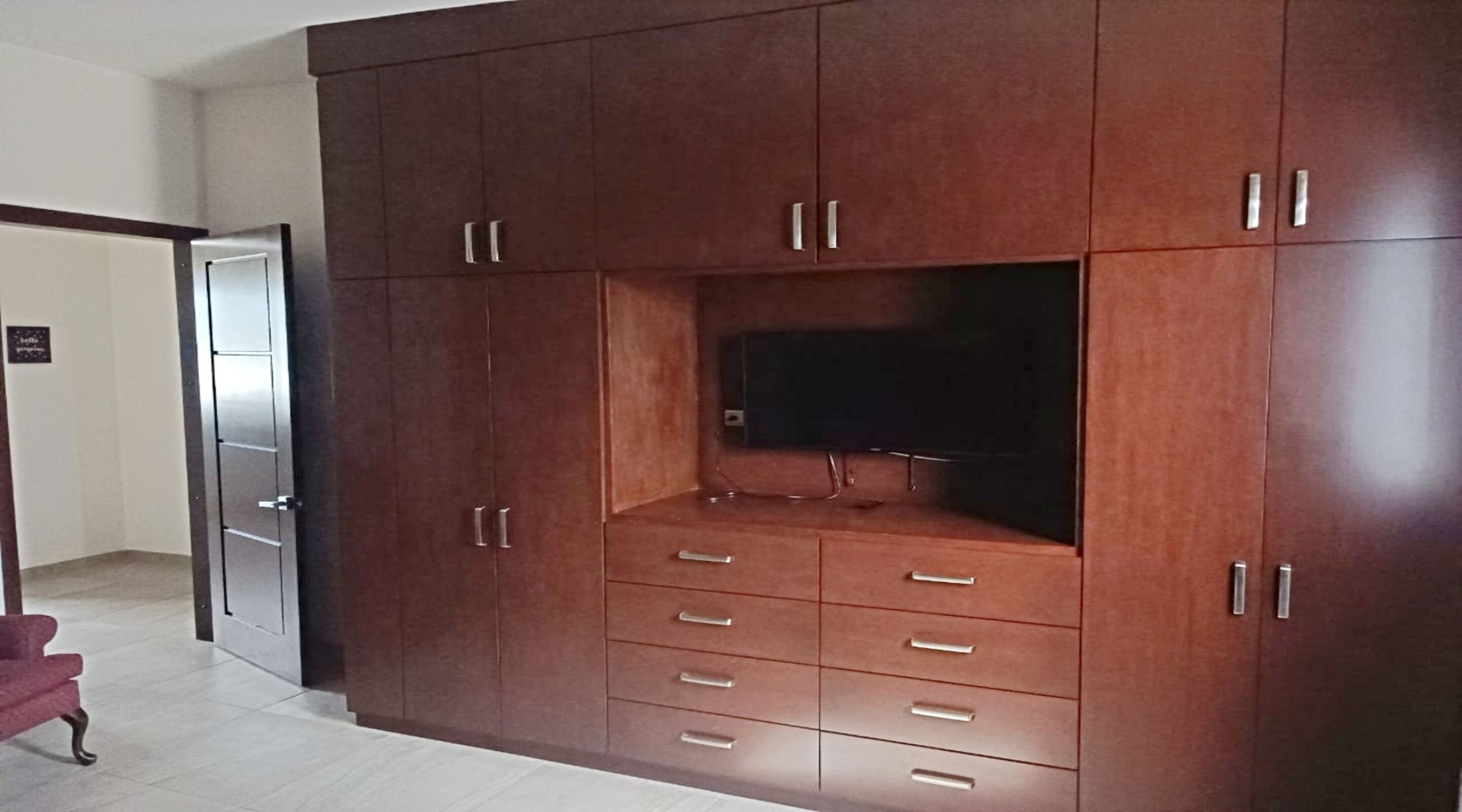A spacious room with a large wooden cabinet featuring multiple drawers and cabinets, a flat-screen TV mounted on the cabinet, and a door leading to another room.
