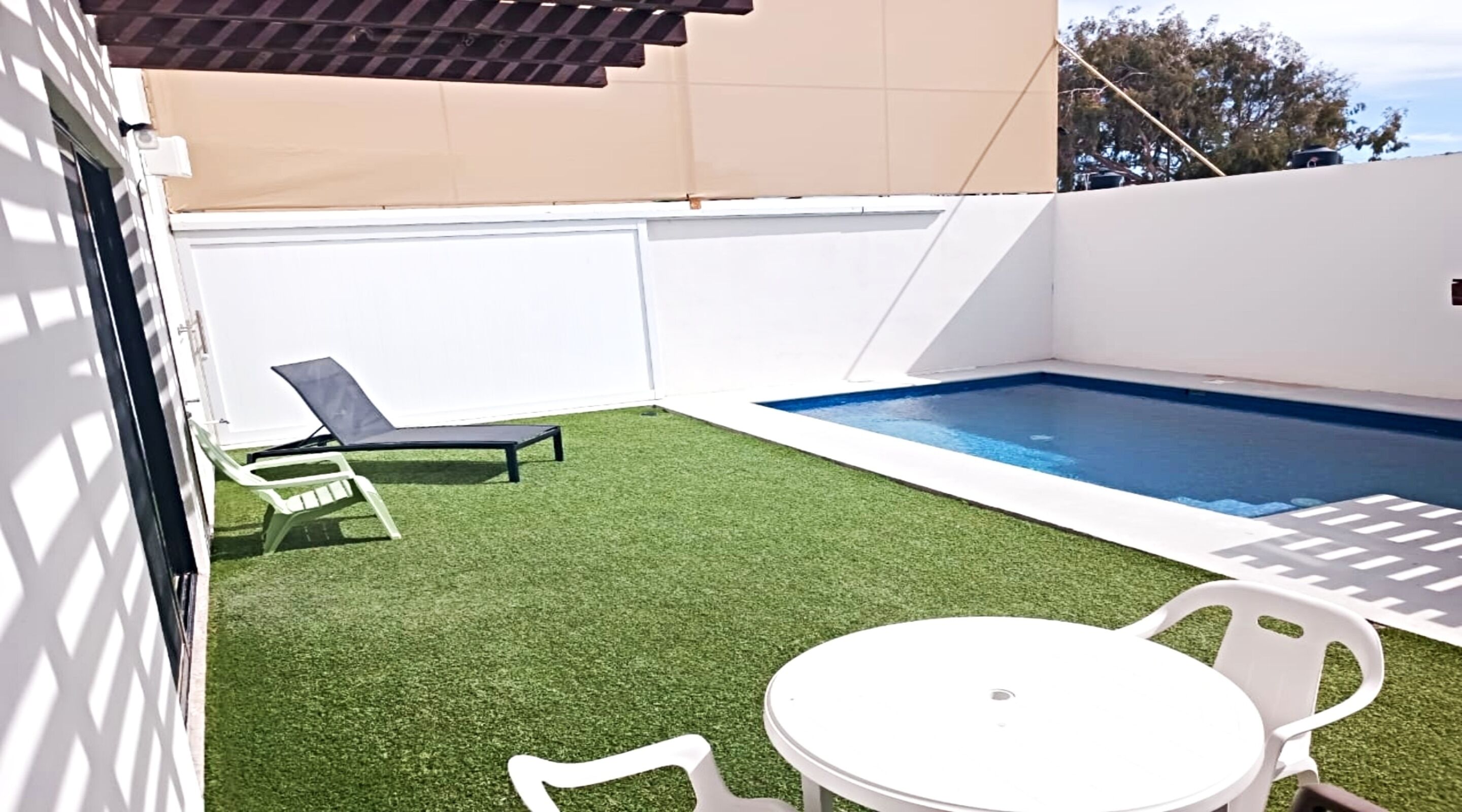 Un pequeño patio trasero con césped artificial, una piscina, una silla de descanso y una mesa con sillas.