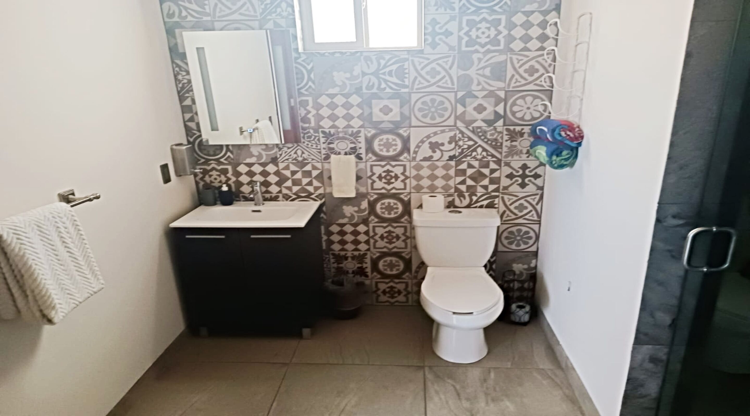 A small bathroom with a white sink, a toilet, and a mirror above the sink. The walls are decorated with patterned tiles.