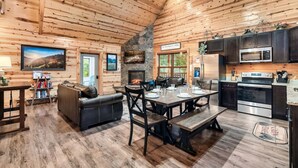 A log cabin interior with a dining area, kitchen, fireplace, and a large window. - Pet Friendly! Private Pool, Mini Golf, Views! (Pigeon Forge)