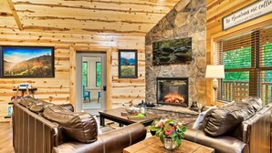 A log cabin interior with a stone fireplace, leather sofas, and a dining table with a vase of flowers. - Pet Friendly! Private Pool, Mini Golf, Views! (Pigeon Forge)