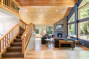 Un vaste espace de vie agrémenté d’un escalier en bois, d’une cheminée en pierre et de grandes fenêtres offrant une vue sur l’extérieur.