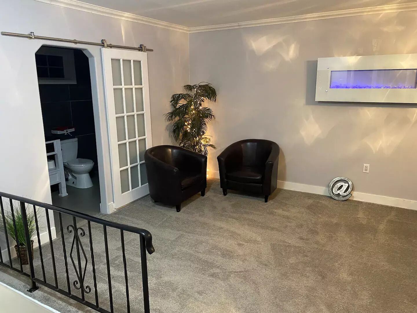 A living room with a grey carpet, two black armchairs, a potted plant, a white door leading to a bathroom, and a mounted TV.